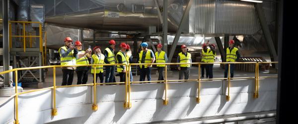 En grupp personer i varselvästar och skyddshjälmar står på en gångbro inne i en avfallsanläggning. Flera fotograferar eller observerar maskinutrustning. Bilden är tagen i samband med VMAB:s deltagande i det internationella initiativet Waste Management for Ukraine (WM4U), där svenska och ukrainska kommunkluster samarbetar för att utveckla avfallshanteringen i Ukraina.